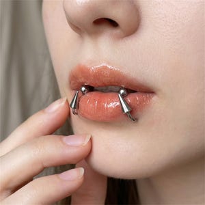 Op de afbeelding: Een zilverkleurig metalen lippiercing sieraad met een gebogen barbell ontwerp. Het sieraad bevat twee gladde bolvormige uiteinden in de mond en twee puntige kegelvormige uiteinden die naar beneden steken vanaf de onderlip.