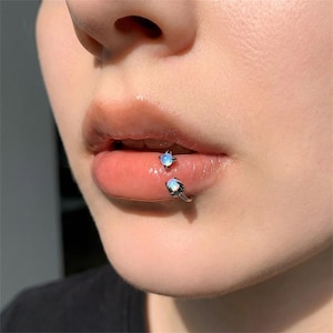 Op de afbeelding: Een zilverkleurige lippiercing met twee kleine, iriserende, ronde cabochonstenen. Het sieraad heeft de vorm van een gebogen staafje dat om de onderlip heen valt, waarbij de ene steen zich in de mond bevindt en de andere aan de buitenkant.