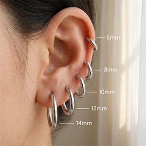 May include: A set of five polished silver-tone hoop earrings of varying sizes, labeled with measurements of 6mm, 8mm, 10mm, 12mm, and 14mm. The hoops are arranged in a vertical row along the ear, showcasing a smooth, rounded metal design.