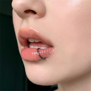 May include: A thin, dark-colored metal lip ring with a textured, hammered surface finish. The circular hoop is positioned securely around the lower lip, resting against the skin. The metal appears to have a matte, oxidized, or dark grey tone.