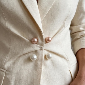 Op de afbeelding: Een beige blazer van gestructureerde stof met een dubbele rij knopen. Het kledingstuk wordt gesloten met twee paar bolvormige knopen, waarbij het bovenste paar een zachte roze tint heeft en het onderste paar een klassieke witte parelmoerafwerking.