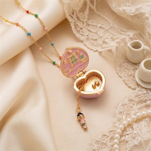 May include: A gold-tone necklace featuring a delicate chain with small multicoloured enamel beads. The pendant is a heart-shaped locket that opens to reveal a miniature room interior with pink walls and a small dangling figurine charm attached.