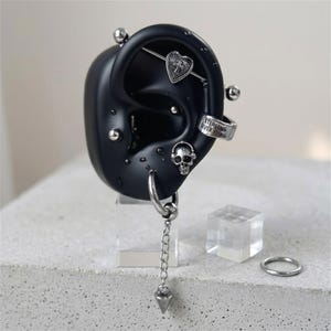 May include: A black ear model displaying multiple silver-toned piercings, including a heart-shaped charm with a cross, a skull-shaped stud, a cuff engraved with the text STERLING SILVER RING, and a dangling chain earring with a pointed tip.