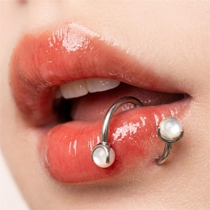 Puede incluir: Una joya corporal de barra circular que presenta un aro de metal plateado pulido con dos cuentas esféricas blancas similares a perlas en cada extremo, colocada a través del labio inferior con una cuenta sobre la piel externa y otra dentro.
