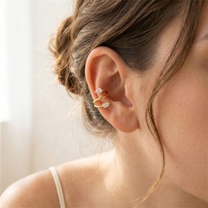 May include: A gold-toned ear cuff featuring a spiral design that wraps around the cartilage. The piece is accented with two clear, pear-shaped stones, one positioned at the top end and one at the bottom end of the spiral metal band.