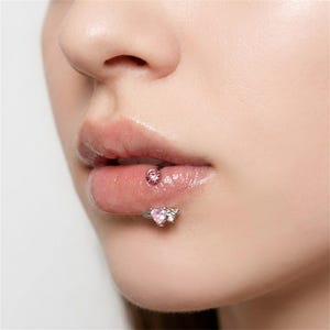 Könnte beinhalten: Ein silberfarbenes Lippenpiercing-Set bestehend aus zwei Teilen. Das obere Teil zeigt einen runden, facettierten rosa Stein, während das untere Teil einen herzförmigen rosa Stein aufweist, der von kleinen metallischen Flügeln flankiert wird.