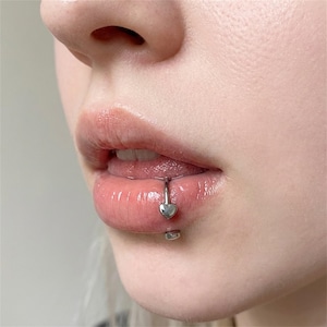 May include: A silver-toned curved barbell lip piercing jewelry featuring a small heart-shaped charm at the top end and a smooth circular bead at the bottom end, positioned vertically through the center of the lower lip.