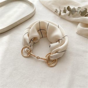 May include: A cream-colored silk scarf featuring a thin black grid pattern, gathered and secured by a polished gold-tone metal horsebit-style clasp. The fabric is soft and draped, with the metallic hardware creating a distinct focal point.