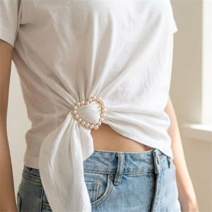 Puede incluir: Una camiseta blanca lisa de manga corta con un accesorio en forma de corazón hecho de pequeñas perlas artificiales redondas de color crema. La tela de la camiseta está fruncida y pasada por el centro del corazón de perlas, creando un efecto anudado.