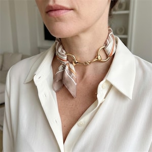 May include: A silk neck scarf featuring a geometric pattern in shades of beige, white, and orange, tied through a polished gold-tone metal horsebit hardware piece. The scarf is worn around the neck, showcasing the metallic detail and fabric.