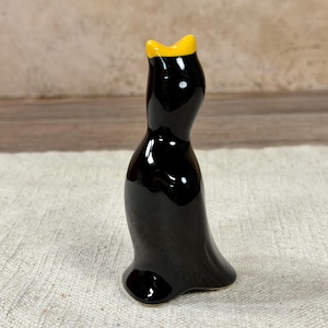 May include: A glossy black ceramic figurine of a penguin with a yellow crown. The penguin has a smooth, curved body and a small base. The figurine is standing upright and appears to be handmade.