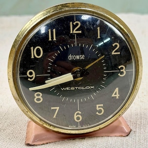 May include: Vintage Westclox alarm clock with a black face, gold-tone bezel, and a pink base. The clock features white numerals, hands, and the word "drowse". The text "WESTCLOX" and "MADE IN U.S.A." are also visible.