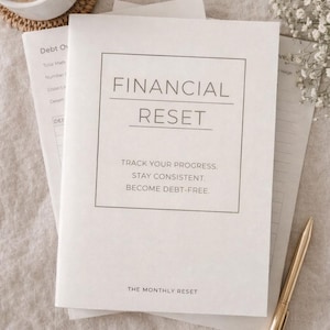 May include: A white notebook titled "FINANCIAL RESET" with text: "TRACK YOUR PROGRESS. STAY CONSISTENT. BECOME DEBT-FREE." A gold pen and a cup of coffee sit nearby. The notebook is part of "THE MONTHLY RESET" collection.
