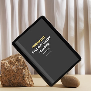 Könnte beinhalten: Ein schwarzes Tablet mit dem Text "MINIMALIST STUDENT TABLET PLANNER" in Gelb und Weiß. Das Tablet wird von einem Felsen und einem kleinen Stein gestützt. Der Hintergrund zeigt eine beige Oberfläche und weiße Vorhänge.