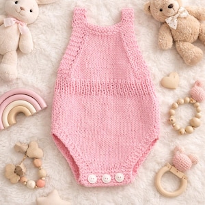Può includere: Un body per neonato rosa chiaro lavorato a mano con tre bottoni bianchi. Il body è circondato da giocattoli per bambini, tra cui un coniglietto di peluche, un orsacchiotto, un arcobaleno di legno e un sonaglio di legno. I giocattoli sono su una superficie bianca e soffice.