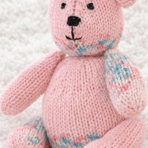 Puede incluir: Un oso de peluche rosa tejido a mano con ojos y nariz bordados en negro. El oso tiene orejas, brazos y piernas pequeños. Las patas y el cuello del oso tienen un diseño estampado en azul y rosa.