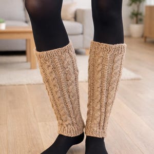 Peut inclure: Une paire de jambières en tricot de couleur beige clair. Les jambières sont portées sur des collants noirs et des chaussettes noires. Le motif tricoté présente un motif torsadé répété. L'arrière-plan montre un sol en bois et un aperçu d'un salon.