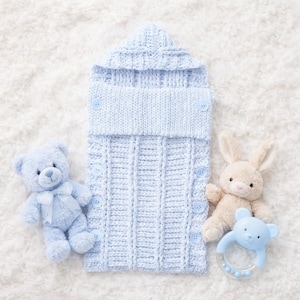 Può includere: Un sacco nanna per neonati in maglia blu chiaro con bottoni, un orsacchiotto abbinato, un coniglietto di peluche beige e un sonaglio blu a forma di orso. Gli oggetti sono disposti su una superficie bianca e soffice.