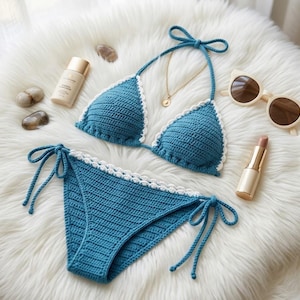 Op de afbeelding: Een handgemaakte blauwe gehaakte bikini met witte rand, bestaande uit een haltertop en een broekje met strik. De bikini wordt getoond met een zonnebril, een gouden ketting en make-up op een zacht witte ondergrond.