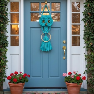 May include: A teal owl-shaped macrame door hanging with a wooden dowel and a large tassel. The door is painted a muted blue and has a brass handle. Two terracotta pots with red flowers flank the door, and a welcome mat is at the base.