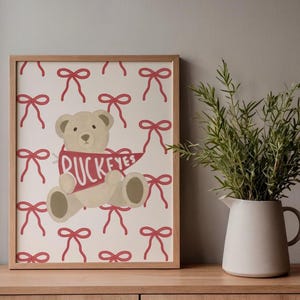 Puede incluir: Un estampado enmarcado con un oso de peluche beige sosteniendo una bandera roja con la palabra "BUCKEYES". El fondo es un patrón de lazos rojos sobre un fondo crema. A la derecha hay una jarra blanca con vegetación.