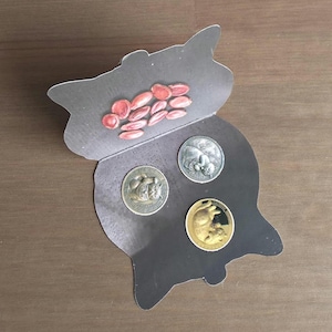 May include: A cat-shaped cardboard display card featuring a printed illustration of pink seeds on the upper flap and three circular coins embedded in the lower section, including one silver-toned, one bronze-toned, and one gold-toned coin with cat motifs.
