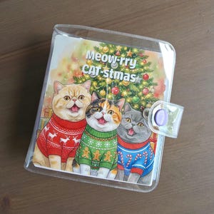 May include: A clear plastic binder featuring an illustration of three cats wearing holiday sweaters in front of a decorated Christmas tree. The text Meow-rry CAT-stmas is printed at the top. The binder has a purple snap button closure.