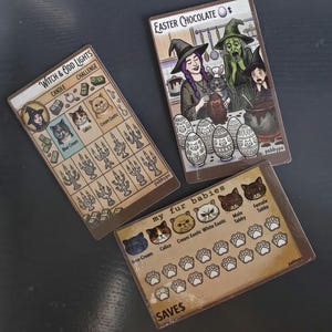 May include: Three rectangular game cards with illustrated themes. One card titled Witch & Odd Lights features cat portraits and candle icons. Another titled Easter Chocolate shows witches and decorated eggs. The third titled my fur babies displays cat faces and paw print trackers.