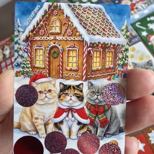 May include: A rectangular sticker sheet featuring a gingerbread house with candy decorations and three cats wearing Christmas attire. The cats are positioned in front of the house, accented by several circular, glittery, and metallic red stickers.