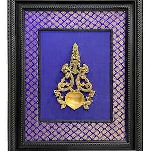 May include: Framed artwork featuring a gold-colored, ornate metal sculpture against a blue background. The sculpture has a heart-shaped base and intricate details. The frame is black with a textured design, and the background fabric has a gold floral pattern.