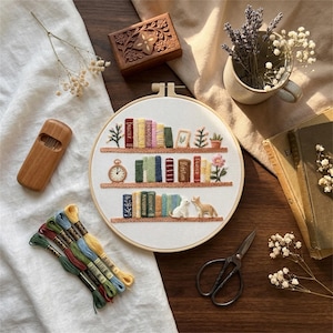 Könnte beinhalten: Ein Stickrahmen mit einem Bücherregal-Design, das mit farbigen Fäden gestickte Bücher, Pflanzen und eine Uhr zeigt. Die Szene umfasst eine Holzbox, eine Schere, Faden und eine kleine Vase mit Lavendel und Blumen. Das Thema ist eine gemütliche, handgefertigte Ästhetik.
