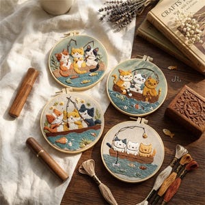 May include: Four embroidered hoops with cat designs, including cats in boats and fishing scenes. The cats are in shades of orange, white, and black. The hoops are displayed on a light surface with craft supplies.