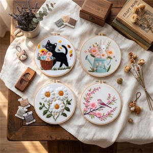 Op de afbeelding: Vier geborduurde hoepels met bloemen- en dierontwerpen. Een zwarte kat met een bloemenmand, een konijn in een gieter, madeliefjes en een vogel op een bloeiende tak worden afgebeeld.