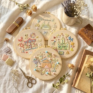 May include: Four embroidered hoops with circus-themed designs, including a carousel, castle, and tent, are displayed. The hoops are beige with colorful thread details. Additional items include a wooden needle case, scissors, and spools of thread.