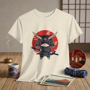 May include: Cream-colored t-shirt featuring a cartoon samurai cat design. The cat is depicted in a black and navy blue samurai outfit with a red sun in the background. The cat is holding a sword. The shirt is on a wooden surface.
