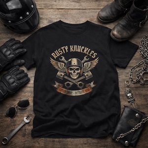 May include: Black t-shirt with a graphic featuring a skull wearing a helmet, wings, and crossed pistons. The text reads "Rusty Knuckles Speed Shop - Detroit, USA". The image includes motorcycle gear like gloves, boots, a helmet, and sunglasses.