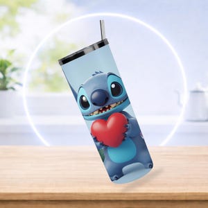 K&ouml;nnte beinhalten: Ein Edelstahlbecher mit einer Cartoonfigur, die ein rotes Herz h&auml;lt. Die Figur ist blau mit gro&szlig;en Augen und einem Grinsen. Der Becher hat einen schwarzen Deckel und einen silbernen Strohhalm.