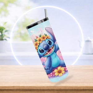 Pode incluir: Caneca azul com um personagem de desenho animado usando uma coroa de flores. A caneca tem um canudo prateado e uma tampa preta. O design apresenta um esquema de cores azul claro, rosa e amarelo. Em uma mesa de madeira.