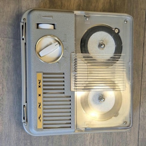 May include: A vintage grey Miny portable reel-to-reel tape recorder featuring a clear plastic cover over two circular reels, a white volume dial, a white control knob, and a gold-colored vertical nameplate with the brand name Miny printed in black.