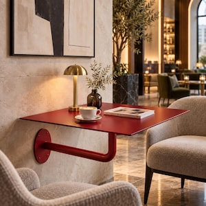 Peut inclure: Une table carrée en métal rouge foncé fixée au mur par un bras de support unique. Sur la table se trouvent une lampe dorée en forme de champignon, un petit vase en verre avec des brins verts, une tasse à café blanche sur soucoupe et un livre.
