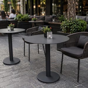 May include: Two round matte black metal bistro tables with central pedestal bases, each paired with a dark brown woven wicker chair featuring a black seat cushion and a small back pillow, arranged on a stone patio with small potted plants.