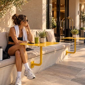 Puede incluir: Una mesa de metal amarillo brillante con un soporte en forma de L montado en un banco de piedra. La superficie cuadrada sostiene un vaso de plástico transparente con un batido verde, una pequeña planta en maceta y un teléfono inteligente negro.