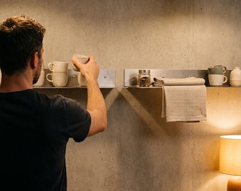 May include: Two long, horizontal, brushed metal wall shelves mounted on a concrete wall. The shelves hold several ceramic mugs, a small glass jar with a metal lid, and a folded beige waffle-textured kitchen towel, positioned above a gas stove.