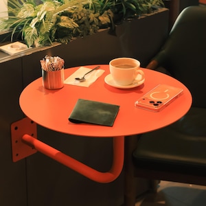 May include: An orange, round side table with a curved support arm. A cup of coffee on a saucer, a spoon, a napkin, a phone, and a dark green wallet are on the table. A metal container holds stir sticks.