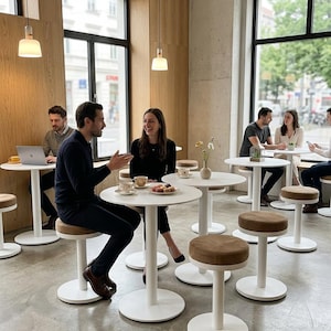 May include: A modern cafe interior featuring several white pedestal tables and matching stools with brown upholstered seats. People are seated at the tables, some working on laptops and others conversing over coffee and small plates of food.
