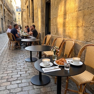 Könnte beinhalten: Ein runder schwarzer Bistro-Tisch aus Metall auf einer Kopfsteinpflasterstraße zeigt zwei weiße Keramiktassen mit Latte-Art-Kaffee, ein goldbraunes Croissant auf einem weißen Teller, ein kleines Glas Wasser und eine kleine Glasvase mit Wildblumen.