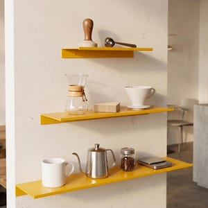 Op de afbeelding: Drie gele wandplanken met koffiezetapparatuur. De bovenste plank bevat een houten stamper en een kleine metalen lepel. De middelste plank heeft een glazen karaf en een witte keramische pour-over. De onderste plank heeft een witte mok, een zilveren waterkoker en een potje koffiebonen.