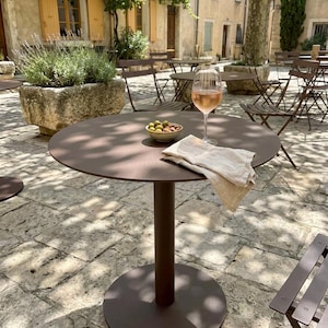 May include: A round, dark brown metal pedestal table featuring a single central column and a circular base. On the tabletop sits a glass of rose wine, a small ceramic bowl filled with green olives and almonds, and a folded beige linen napkin.