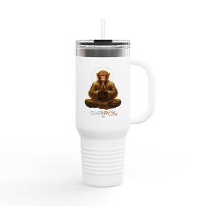 May include: White travel mug with a handle and a straw. The mug features a brown monkey in a meditative pose with a gold halo. The text "SHRP Clo" is printed below the monkey.