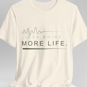 Könnte beinhalten: Ein cremefarbenes Kurzarm-T-Shirt mit einer Grafik einer salbeigrünen Schallwellenlinie über dem Text "LESS NOISE." und "MORE LIFE." in dunkelgrünen Großbuchstaben, wobei eine horizontale Linie den letzten Satz unterstreicht.